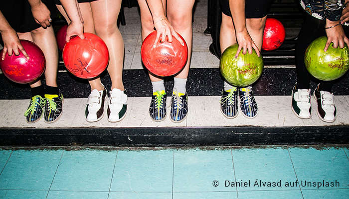 Von 5 Personen sieht man neben einer Bowling Bahn die Bowlingschuhe sowie die Hände, die jeweils eine Bowling-Kugel halten.