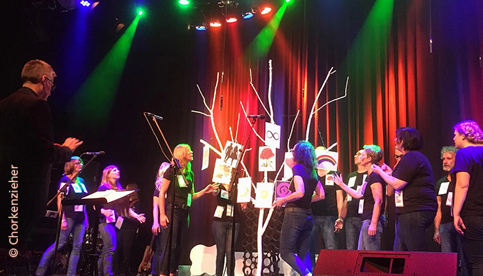 Der Chor "Chorkenzieher" singt auf der Bühne. Die Beleuchtung ist farbenfroh.