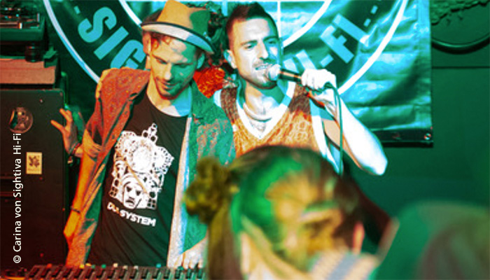 Zwei junge Männer am Soundsystem im Vordergrund Partypeople im Hintergrund Projektion des Schriftzugs Sightiva Hi-Fi